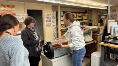 BPS print shop operator Victoria Hendricks describes how to run the laminator in district's Print Shop with student Charlie Bonstein and job coach paraeducator Karen Morris listening.